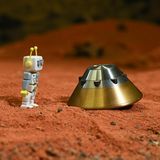 Ein Spielzeug-Marsmensch steht auf rotem Untergrund und blickt auf eine  Testkapsel des ExoMars-Landemoduls.