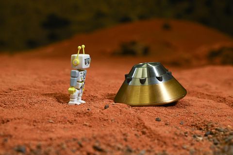 Ein Spielzeug-Marsmensch steht auf rotem Untergrund und blickt auf eine  Testkapsel des ExoMars-Landemoduls.