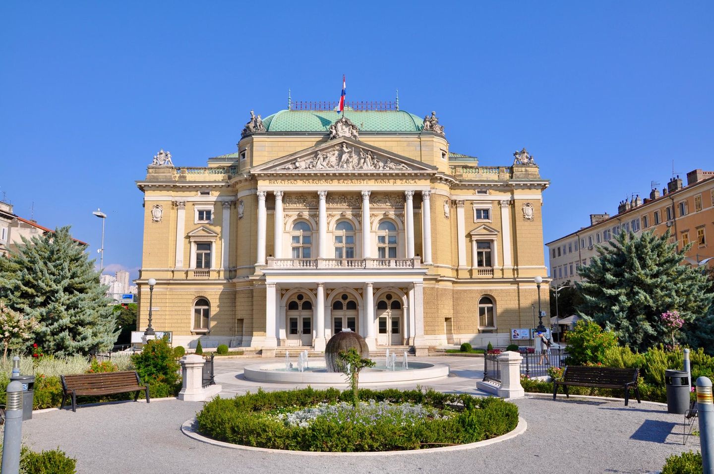 Nationaltheater mit Säulen und Fahne, davor angelegter Garten mit Bänken