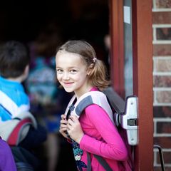 Ein Mädchen mit Schulranzen steht am Schuleingang und lächelt in die Kamera